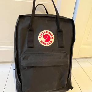 Svart ryggsäck från Fjällräven - Säljer en klassisk svart Fjällräven Kånken ryggsäck. Den har justerbara axelremmar och ett stort huvudfack med dragkedja samt ett smalare fack baktill med dragkedja. Framtill finns en mindre ficka med dragkedja och på sidorna praktiska öppna fickor. Perfekt för vardagsbruk och utflykter. Modell 27171 Kånken 13. Nypris 1199 kr.