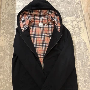 Svart hoodie från Burberry - Säljer en stilren svart hoodie från Burberry med klassiskt rutigt foder i huvan. Jackan har dragkedja framtill och en broderad logotyp på bröstet. Perfekt för en trendig look.