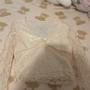 Beige spetsblus med rosett - Säljer en elegant beige spetsblus med långärmade ärmar och en söt rosett framtill. Blusen har ett vackert blommönster och en v-ringad design som ger en romantisk touch.