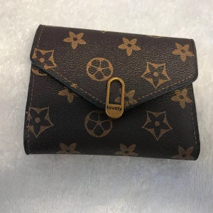 Brun mönstrad plånbok från Lovely - Snygg brun plånbok med klassiskt mönster från Lovely. Den har ett praktiskt fack för kort och en säker stängning med gulddetalj. Perfekt för att hålla ordning på dina kort och kontanter. 95kr/st . Helt ny 