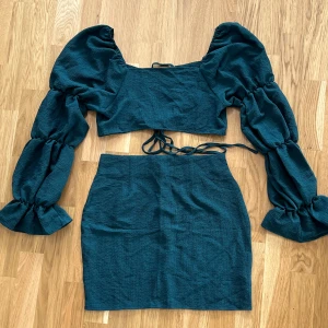 Mörkgrön tvådelad klänning - Trendig mörkgrön tvådelad klänning med en tight passform. Toppen har långa puffärmar och en knytning framtill som ger en snygg detalj. Kjolen är kort och figurnära, perfekt för en stilren look.