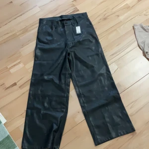 Svarta byxor i syntetmaterial - Snygga svarta byxor i syntetmaterial med hög midja och vida ben. Byxorna har en dragkedja framtill och bälteshällor för extra stil. Perfekta för en trendig look.