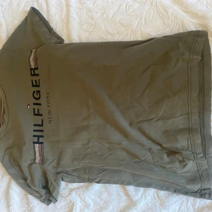 Olivgrön t-shirt från Tommy Hilfiger - Säljer en olivgrön t-shirt från Tommy Hilfiger med tryck på framsidan. T-shirten har korta ärmar och en klassisk rund hals. Perfekt för en avslappnad stil.