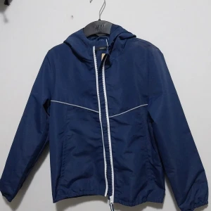 Blå vindjacka  - Säljer en snygg blå vindjacka från Name it  i storlek EU 140. Jackan har en vit dragkedja och en praktisk huva. Perfekt för blåsiga dagar och har en stilren design med reflekterande detaljer.