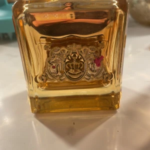 Juicy Couture Viva La Juicy Gold Couture Eau De Parfum 100 ml - Juicy Couture Viva La Juicy Gold Couture är en lyxig parfym i en elegant glasflaska med gulddetaljer och en rosa rosett. Flaskan rymmer 100 ml av en doft som utstrålar glamour och elegans. Perfekt för den som vill ha en doft med en touch av lyx. Locket är tyvärr borta darför sänkte jag priset ganska mycket från 1200 till 650