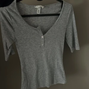 Grå ribbad topp från H&M - Säljer en stilren grå ribbad topp från H&M med trekvartsärm och knappar framtill. Perfekt för en avslappnad look. Passar bra till jeans eller kjol.