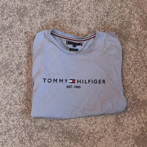 Ljusblå t-shirt från Tommy Hilfiger - Säljer en ljusblå t-shirt från Tommy Hilfiger med tryck 'Est. 1985' på framsidan. T-shirten är kortärmad och har en klassisk rund halsringning. Perfekt för en avslappnad stil. Har ett litet hål på ryggen, annars är den nästan oanvänd.