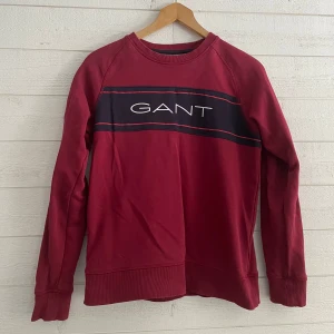 Röd barn sweatshirt från GANT - Snygg röd sweatshirt från GANT med ett svart och vitt band över bröstet där märket är tryckt. Tröjan har långa ärmar och ribbade muddar vid ärmslut och nederkant. Perfekt för en avslappnad stil. Storlek 170.