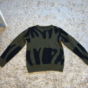 Olivgrön och svart mönstrad tröja - Säljer en olivgrön tröja med svart mönster. Tröjan har långa ärmar och en rund halsringning. Perfekt för en unik och stilren look.