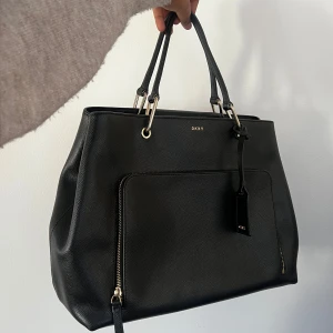 DKNY väska - Säljer en DKNY väska som är mycket bra skick. Använt fåtal gånger. Den är väldigt rymlig och har flera användbara fack så passar perfekt som jobb/skol väska med laptop/böcker i och handväska när man ska åka bort etc.   Måtten är 24cm höjd, 36cm bredd, 17cm djup. Ett axelband ingår som går att sätta på/ta av vid behov.   Väskan kan fraktas och pris kan absolut diskuteras vid snabb affär. 
