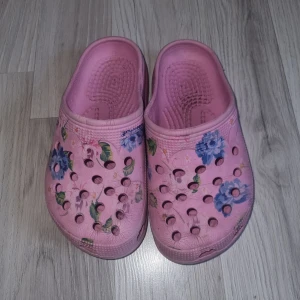 Rosa blommiga tofflor/sandaler  - Säljer ett par rosa tofflor/sandaler med blommigt mönster. De har en bekväm passform med perforerade detaljer för ventilation. Perfekta för sommarens varma dagar.