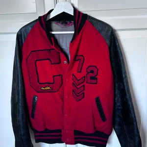 Röd och svart baseball jacka med bokstavsdetaljer - Snygg röd och svart baseball jacka med stora bokstäver och siffror på framsidan. Jackan har svarta ärmar och ribbade muddar i svart och rött. Perfekt för en cool och avslappnad stil. Hör av er vid intresse/ frågor!
