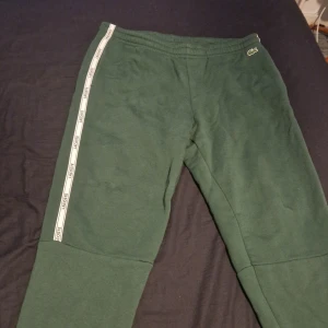 Gröna mjukisbyxor från Lacoste - Snygga gröna mjukisbyxor från Lacoste med en vit rand längs sidan där Lacoste-loggan är tryckt. Byxorna har en avslappnad passform och är perfekta för en bekväm stil. Den ikoniska krokodilloggan är broderad på framsidan. Använt 1-2 gånger.