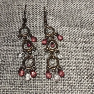 Örhängen med pärlor i brons och rosa - Snygga örhängen i brons med droppformade detaljer och hängande pärlor i rosa och klara nyanser. Perfekta för att ge en bohemisk touch till din stil. De har en vintagekänsla med sina intrikata mönster och färgkombinationer. Det saknas en rosa pärla.