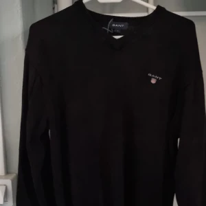 Svart tröja från GANT - Säljer en stilren svart tröja från GANT med v-ringning och broderad logotyp på bröstet. Tröjan har långa ärmar och ribbade muddar vid ärmslut och nederkant. Perfekt för en klassisk look.