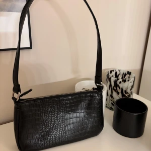 Svart axelväska - Snygg svart axelväska i syntet med krokodilmönster. Väskan har en praktisk dragkedja upptill och en justerbar axelrem. Rätt rymlig för att vara den typen av väska! Köpt på Ullared för 199kr ⭐️👜Inte använd särskilt många gånger, den är välbehållen! 