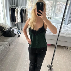 Grön sammetlinne med spetsdetaljer - Snyggt grönt sammetlinne med svarta spetsdetaljer vid halsringning och nederkant. Linnet har smala axelband och en lös passform, perfekt för en elegant look. Märket är H&M. Hittar inte storleken men den passar mig som vanligen har XS eller S. Passar nog S-L beroende på hur tight man vill att den ska sitta