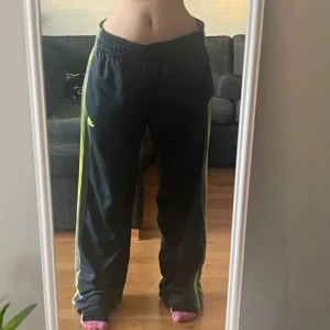 Svarta träningsbyxor från Adidas - Säljer ett par svarta träningsbyxor från Adidas med neongula ränder längs sidorna. Byxorna har en elastisk midja och är perfekta för sportiga aktiviteter. Adidas-loggan syns tydligt på sidan.