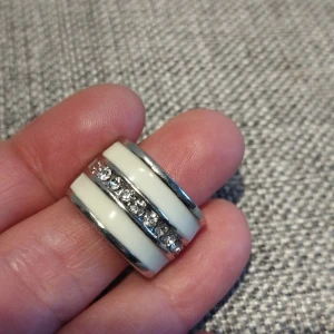 Vit och silverfärgad ring med stenar storlek 8 - Elegant ring i vit och silverfärg med en rad av glittrande stenar. Ringen har en bred design och en modern look, perfekt för att ge en extra touch till din stil.