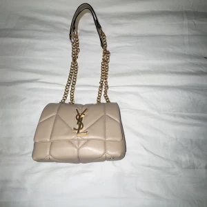 Beige axelväska från Yves Saint Laurent - Snygg beige axelväska från Yves Saint Laurent med quiltad design och guldfärgad kedja. Väskan har det ikoniska YSL-emblemet på framsidan och är tillverkad i skinn. Perfekt för att ge en lyxig touch till din outfit.