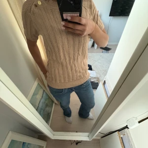Beige stickad tröja med knappar. - Säljer en snygg beige stickad tröja med korta ärmar och dekorativa knappar vid axeln. Tröjan har ett ribbat mönster och är perfekt.Passar bra till jeans eller kjol. Perfekt för vår och höst! 