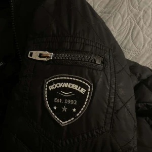 Svart dunjacka från Rockandblue - Snygg svart dunjacka från Rockandblue med quiltade detaljer och en avtagbar huva med pälsimitation. Jackan har dragkedja och knappar framtill samt flera praktiska fickor med dragkedjor. Perfekt för kyliga dagar!