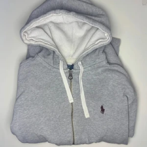 Grå hoodie från Ralph Lauren /Priset kan diskuteras 😁 - Säljer en klassisk grå hoodie från Ralph Lauren med dragkedja och vit snörning. Den har en broderad logotyp på bröstet och är tillverkad i bomull och polyester. Perfekt för en avslappnad stil.    (Du kommer absolut kommer inte ångra ditt köp!)