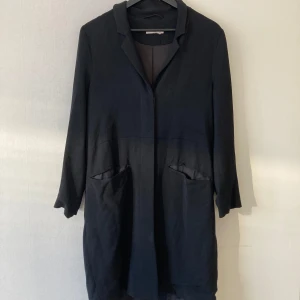 Svart kappa med knappar - Kappa/trenchcoat i rakpassform från märket Wera. Fickor fram och dekorativa knappar på ärmarna. Stl 36