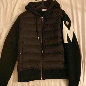 Svart cardigan från Moncler den är helt ny - Snygg svart cardigan från Moncler med dragkedja och huva. Jackan har ett stort vitt M på ärmen och Moncler-logga. Perfekt för kyliga dagar med stil.