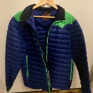 Blå och grön pufferjacka - Säljer en blå pufferjacka med gröna detaljer och dragkedja. Jackan har en hög krage och är perfekt för kyligare dagar. Den har två fickor med dragkedjor och en quiltad design som ger extra värme. Diskutera om priset där vi båda är glada