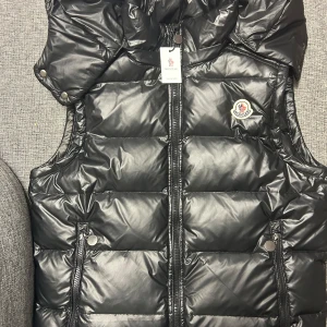 Svart dunväst från Moncler - Snygg svart dunväst från Moncler med dragkedja och tryckknappar. Västen har en quiltad design och en hög krage för extra värme. Perfekt för kyligare dagar. Storlek: XS Passat mig som har storlek S