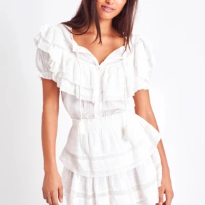 Vit studentklänning  - Charmig vit klänning med volanger och knytband framtill. Den har korta ärmar och en luftig passform, perfekt för varma dagar. Denna klänning har en romantisk känsla med sina lager av volanger och mjuka detaljer.