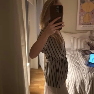 Randig blus med definerad midja - Säljer en stilren randig blus med knytband i midjan. Blusen har korta ärmar och ett omlottmönster framtill. Perfekt för en chic och avslappnad look.