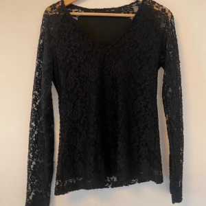 Svart spetsblus - Elegant svart spetsblus med långärmad design och v-ringad hals. Perfekt för en stilfull look med sitt genombrutna spetsmönster.