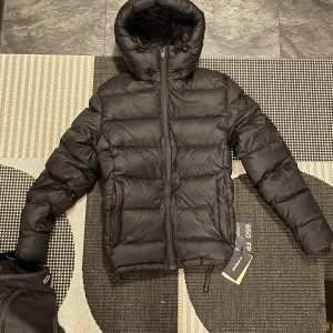 Svart dunjacka med huva från Everest - Säljer en svart dunjacka med huva och dragkedja. Jackan är quiltad och har en pufferdesign som ger en skön och varm känsla. Perfekt för kyligare dagar. 🖤storlek M, nypris 2500kr