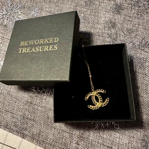 Guldigt halsband med logotyp cc chanel från reworked treasure  - Elegant halsband med en guldpläterad logotypberlock. Kedjan är tunn och smidig, vilket ger en stilren look. Perfekt för att lägga till en touch av lyx till din outfit. Smycke från reworked treasures. Cc Chanel.