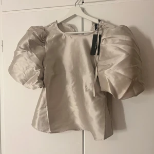 Beige blus med puffärmar från vero mosa - Elegant beige blus från vero Moda med stora puffärmar och knytband baktill. Tillverkad i ett glansigt material som ger en lyxig känsla. Perfekt för att lyfta vilken outfit som helst. Champanj/beige färg. Helt ny