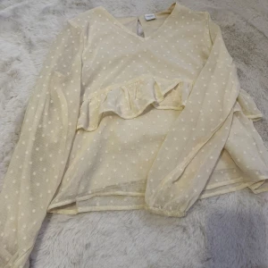 Krämvit/beige prickig blus från Lindex kids - Charmig beige blus från Lindex med vita prickar och volangdetalj framtill. Blusen har en v-ringad hals och långa ärmar, perfekt för en stilren look. Passar utmärkt för både vardag och speciella tillfällen. Den är i storlek 134 och mycket bra skick bortsett från att den har en liten fläck vid ärmen som visas på en av bilderna men den är inte så märkbar. 