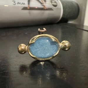  ring med gulddetaljer - Unik ring med en stor blå glittrig sten i mitten, omgiven av guldfärgade detaljer. Ringen har en elegant och iögonfallande design som passar perfekt för speciella tillfällen.