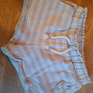 Randiga shorts från Zara - Säljer ett par bekväma och stilrena randiga shorts från Zara. De har en elastisk midja med snörning och är perfekta för en avslappnad look. Shortsen är i en ljus färgskala med vita och beige ränder.