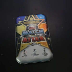 Match Attax samlarkort i plåtlåda - Säljer en plåtlåda med Match Attax samlarkort från säsongen 2021/22. Lådan har en färgglad design med stjärnor och logotyper för Europa League, Champions League och Europa Conference League. Perfekt för fotbollsfans och samlare!