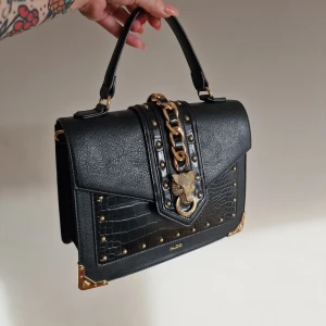 Svart handväska från Aldo - Snygg svart handväska från Aldo med gulddetaljer och kedja. Väskan har en strukturerad yta med krokodilmönster och dekorativa nitar. 