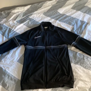 Svart träningfleece från Nike - Snygg svart träningsfleece från Nike med vita detaljer och dragkedja framtill. Fleecen har långa ärmar och en hög krage. Perfekt för sportiga aktiviteter eller en avslappnad stil.