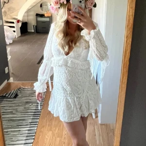 Vit spetsklänning med volangdetaljer - Säljer en romantisk vit spetsklänning med v-ringning och volangdetaljer. Klänningen har långa ärmar och en kort längd, perfekt för sommarens fester. Den har en vacker struktur och ger en elegant look. Perfekt för studenten! Aldrig använd!! Liten i storlek.💕💕