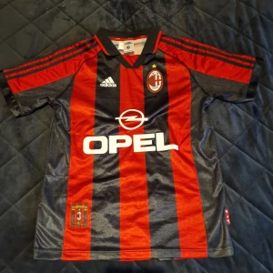 AC Milan fotbollströja med Ronaldinho 80 - Säljer en klassisk AC Milan fotbollströja från Adidas med Ronaldinho och nummer 80 på ryggen. Tröjan är randig i rött och svart med korta ärmar och Opel som sponsorlogga på framsidan. Perfekt för samlare eller fans av laget!
