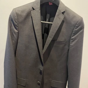 Grå ny kostym med byxor från J. Lindeberg nypris 4499 - Snygg grå kostym från J. Lindeberg, perfekt för formella tillfällen. Kavajen har en klassisk design med två knappar och fickor fram. Byxorna matchar i samma grå nyans och har en stilren passform. Tillverkad i Biella, vilket garanterar hög kvalitet.