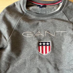 Grå sweatshirt från GANT - Säljer en stilren grå sweatshirt från GANT med broderad logga och ett emblem på bröstet. Tröjan har en klassisk rund hals och långa ärmar. Perfekt för en avslappnad stil.