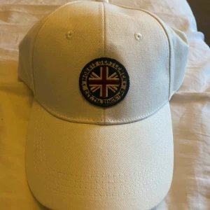 Vit keps från Morris  - Snygg vit Morris keps med en broderad detalj framtill som har en brittisk flagga och text. Perfekt för en avslappnad stil och skydd mot solen. Justerbar passform för bekvämlighet.