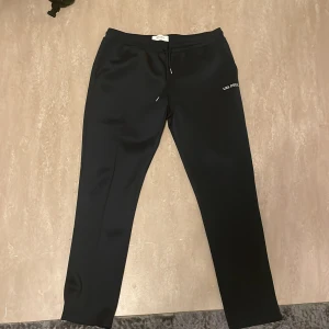 Svarta/mörk blåa byxor från Les Deux - Snygga svarta byxor från Les Deux med broderad logga. De har en stilren design med snörning i midjan för en bekväm passform. Perfekta för en avslappnad men stilfull look.