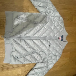 Ljusgrå Lacoste Jacka - Säljer en ljusgrå jacka från Lacoste. Jackan har en quiltad design och är utrustad med dragkedja framtill. Den har långa ärmar och två praktiska fickor med dragkedja. Perfekt för kyligare dagar.